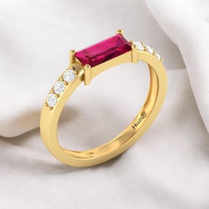 Puede incluir: Un anillo dorado con una piedra preciosa roja rectangular y vibrante como pieza central. El anillo está adornado con pequeñas piedras preciosas redondas y transparentes a lo largo de la banda. La palabra "Huzurr" está grabada en el interior del anillo.