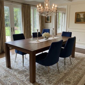 May include: A dining room set with a dark wood table and six navy blue chairs. A crystal chandelier hangs above the table, and a floral rug is beneath. The room has large windows with beige curtains.