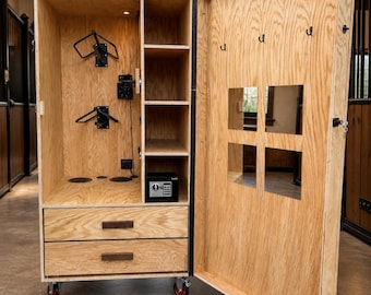 Puede incluir: Un baúl de madera para equipo ecuestre con una puerta abierta, que revela estantes, cajones y una pequeña caja fuerte. El interior incluye soportes para sillas de montar y ganchos. El baúl está sobre ruedas con detalles en rojo.