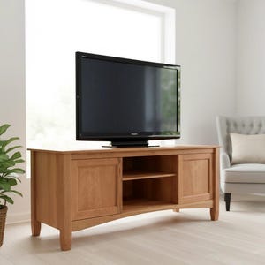 May include: A light brown wooden TV stand with two doors and an open shelf in the middle. A black flat-screen TV sits on top. The stand has a simple, classic design and is placed in a bright room with a window.