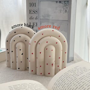 May include: Two ceramic bookends shaped like arches with a white base and red or black hearts. The bookends are on a white surface with a book in front of them.