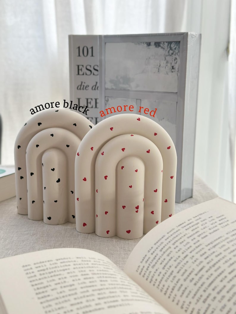 May include: Two ceramic bookends shaped like arches with a white base and red or black hearts. The bookends are on a white surface with a book in front of them.