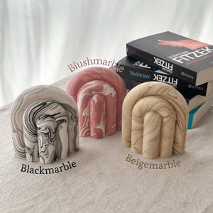 May include: Three decorative bookends in the shape of an arch with a marbled design. The bookends are in black and white, pink and white, and beige and white.