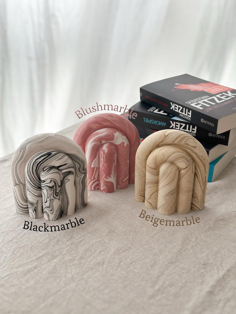 May include: Three decorative bookends in the shape of an arch with a marbled design. The bookends are in black and white, pink and white, and beige and white.