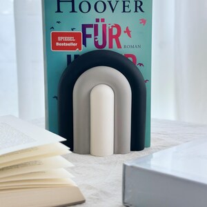 May include: A black, gray, and white rainbow-shaped bookend holding up a book titled "F&uuml;r Immer" by Colleen Hoover. The book has a red and white sticker that says "Spiegel Bestseller" and the word "Roman".