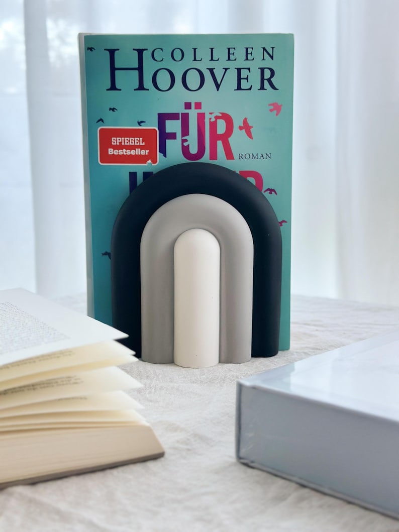 May include: A black, gray, and white rainbow-shaped bookend holding up a book titled "F&uuml;r Immer" by Colleen Hoover. The book has a red and white sticker that says "Spiegel Bestseller" and the word "Roman".
