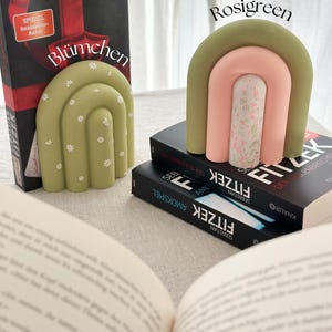 May include: Two green and pink ceramic bookends shaped like arches with a floral pattern. The bookends are on top of two books with the title "Fitzek" on the cover.
