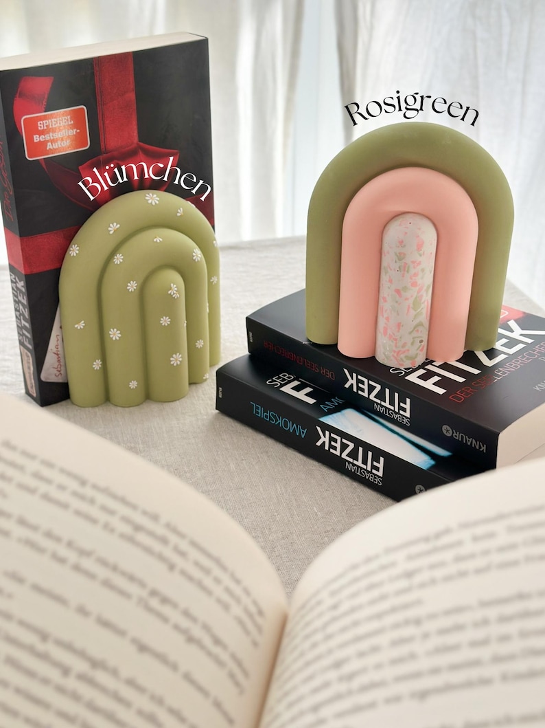 May include: Two green and pink ceramic bookends shaped like arches with a floral pattern. The bookends are on top of two books with the title "Fitzek" on the cover.