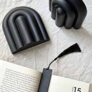 May include: Two black bookends shaped like abstract arches. The bookends are on a white surface. A black bookmark with a black tassel is on top of a book with the text "KAPITEL 15" on the page.