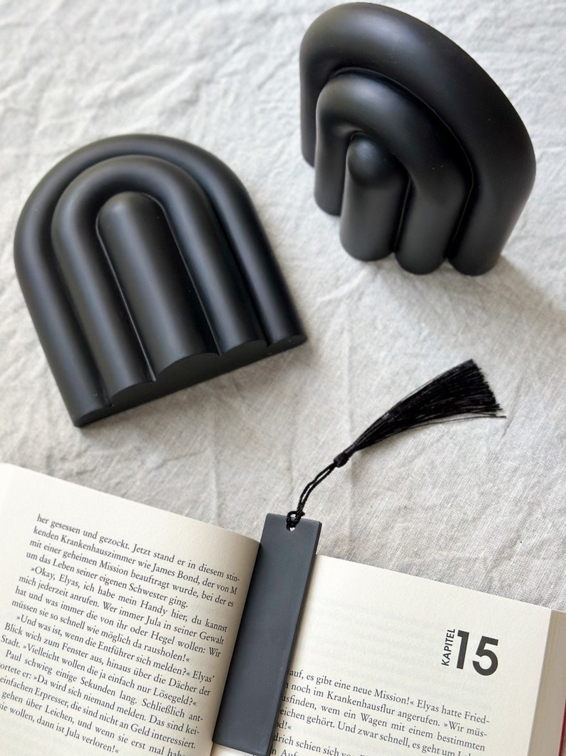May include: Two black bookends shaped like abstract arches. The bookends are on a white surface. A black bookmark with a black tassel is on top of a book with the text "KAPITEL 15" on the page.