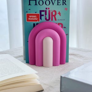 May include: A pink and white bookend shaped like an arch. The bookend is holding up a book titled "F&uuml;r" by Colleen Hoover.