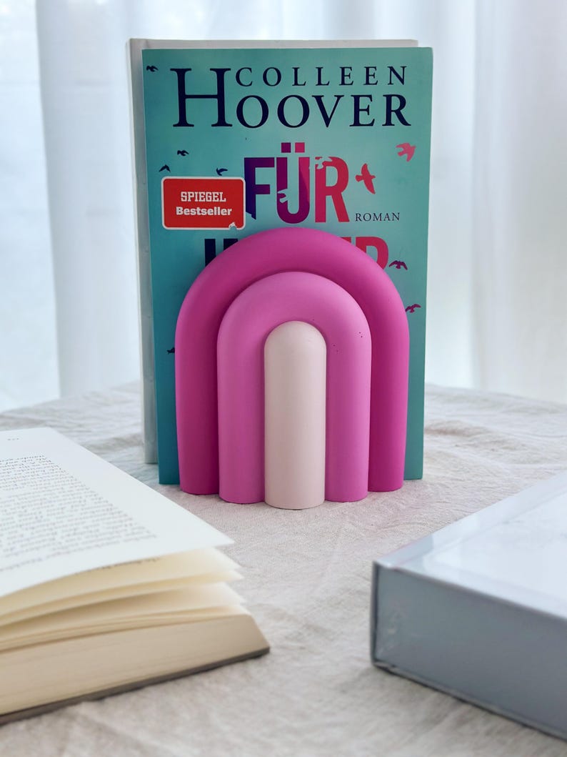 May include: A pink and white bookend shaped like an arch. The bookend is holding up a book titled "F&uuml;r" by Colleen Hoover.