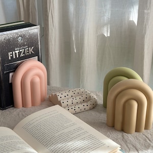 May include: Three decorative bookends in the shape of an arch. The bookends are made of a smooth, solid material and are painted in pastel colors: pink, green, and beige. The bookends are sitting on a white surface with a book open in front of them.