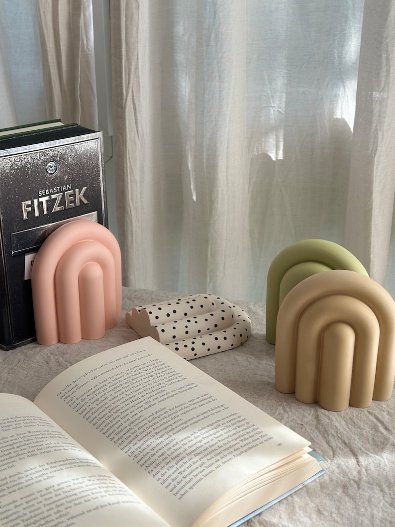 May include: Three decorative bookends in the shape of an arch. The bookends are made of a smooth, solid material and are painted in pastel colors: pink, green, and beige. The bookends are sitting on a white surface with a book open in front of them.