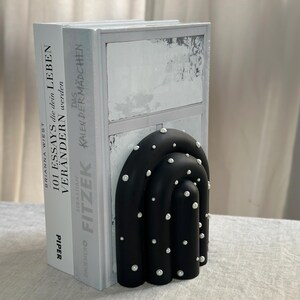 May include: Black bookends shaped like an arch with white pearl-like accents. The bookends are holding up two books with white covers. The book on the left has the title "101 Essays that will change your life" and the book on the right has the title "The Calendar Girl".