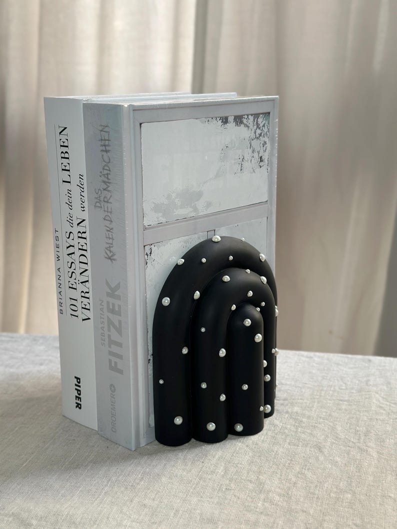 May include: Black bookends shaped like an arch with white pearl-like accents. The bookends are holding up two books with white covers. The book on the left has the title "101 Essays that will change your life" and the book on the right has the title "The Calendar Girl".