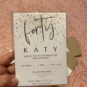 Puede incluir: Una tarjeta de invitaci&oacute;n blanca con detalles de purpurina dorada. El texto de la tarjeta dice "forty" en letra cursiva, seguido de "KATY" en may&uacute;sculas. La invitaci&oacute;n es para la fiesta de cumplea&ntilde;os n&uacute;mero 40 de Katy, que se celebrar&aacute; el s&aacute;bado 6 de mayo a las 7:30 PM en The Engine Shed. RSVP a Jamie.