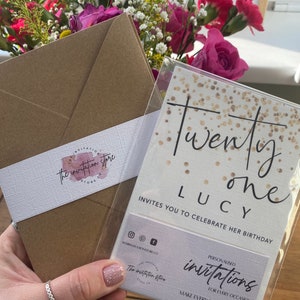 Puede incluir: Un sobre marr&oacute;n con una etiqueta blanca que dice "the invitation store" y una tarjeta dorada con purpurina con el texto "Twenty one Lucy invites you to celebrate her birthday" y una tarjeta blanca m&aacute;s peque&ntilde;a que dice "Invitaciones personalizadas para cada ocasi&oacute;n haz que cada evento sea personal".