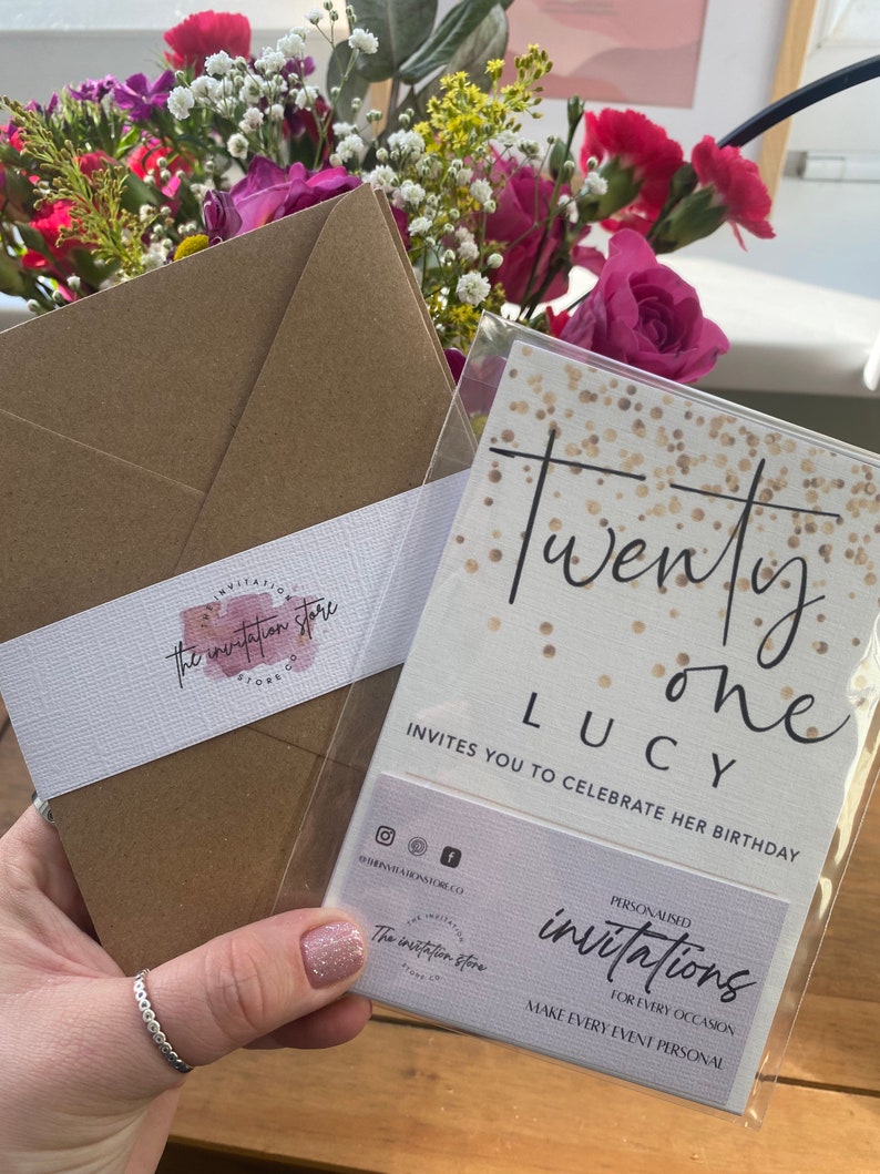 Puede incluir: Un sobre marr&oacute;n con una etiqueta blanca que dice "the invitation store" y una tarjeta dorada con purpurina con el texto "Twenty one Lucy invites you to celebrate her birthday" y una tarjeta blanca m&aacute;s peque&ntilde;a que dice "Invitaciones personalizadas para cada ocasi&oacute;n haz que cada evento sea personal".