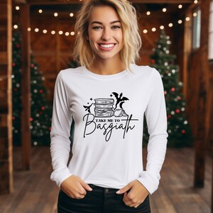 Puede incluir: Camiseta blanca de manga larga con un diseño gráfico negro. El diseño incluye una pila de libros, murciélagos y el texto "TAKE ME TO Basgiath". La camiseta es de cuello redondo.