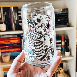 Peut inclure: Un gobelet en verre transparent avec un squelette tenant une pile de livres et des motifs floraux. Le squelette est blanc et les fleurs sont roses et bleues.