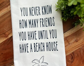 May include: White kitchen towel with black text that reads "You never know how many friends you have until you have a beach house". A black and white starfish is printed below the text.