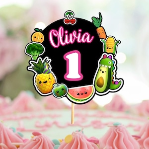 May include: A black and white cake topper with a pink circle that says "Olivia 1" surrounded by cartoon fruit and vegetables. The cake topper is on a pink and white cake with colorful sprinkles.
