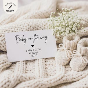 May include: A white rectangular announcement card with the black text Baby on the way, a small heart icon, and the text BABY SMITH AUGUST 2026. The card is placed next to a pair of cream-colored knit baby booties and a small bunch of white flowers.