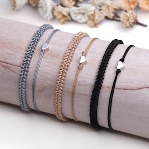 Könnte beinhalten: Eine Auswahl handgefertigter Armbänder in verschiedenen Farben, darunter Grau, Beige und Schwarz. Jedes Armband hat einen kleinen silbernen Herzanhänger. Die Armbänder werden auf einer Holzoberfläche präsentiert, die ihre unterschiedlichen Designs und Texturen hervorhebt.