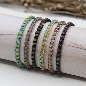 Könnte beinhalten: Ein Set aus sieben bunten Perlenarmbändern, jedes mit einer anderen Farbe und einem anderen Muster. Die Armbänder bestehen aus kleinen, quadratischen Perlen, die auf einer Schnur aufgereiht sind. Die Armbänder sind in einer Reihe angeordnet, wobei die hellste Farbe oben und die dunkelste Farbe unten ist.
