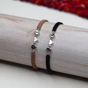 Könnte beinhalten: Zwei Freundschaftsarmbänder mit schwarzen und braunen Schnüren, silbernen Herzanhängern und silbernen Buchstabenperlen. Die Armbänder buchstabieren "J" und "K".