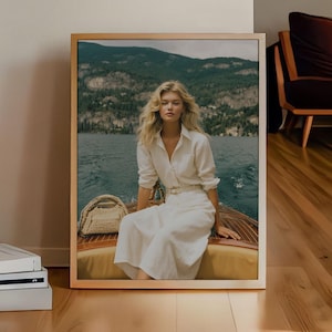 Puede incluir: Una fotografía enmarcada de una mujer vestida de blanco sentada en un barco. La foto está en un marco de madera clara y está apoyada contra una pared. El suelo es de madera noble.
