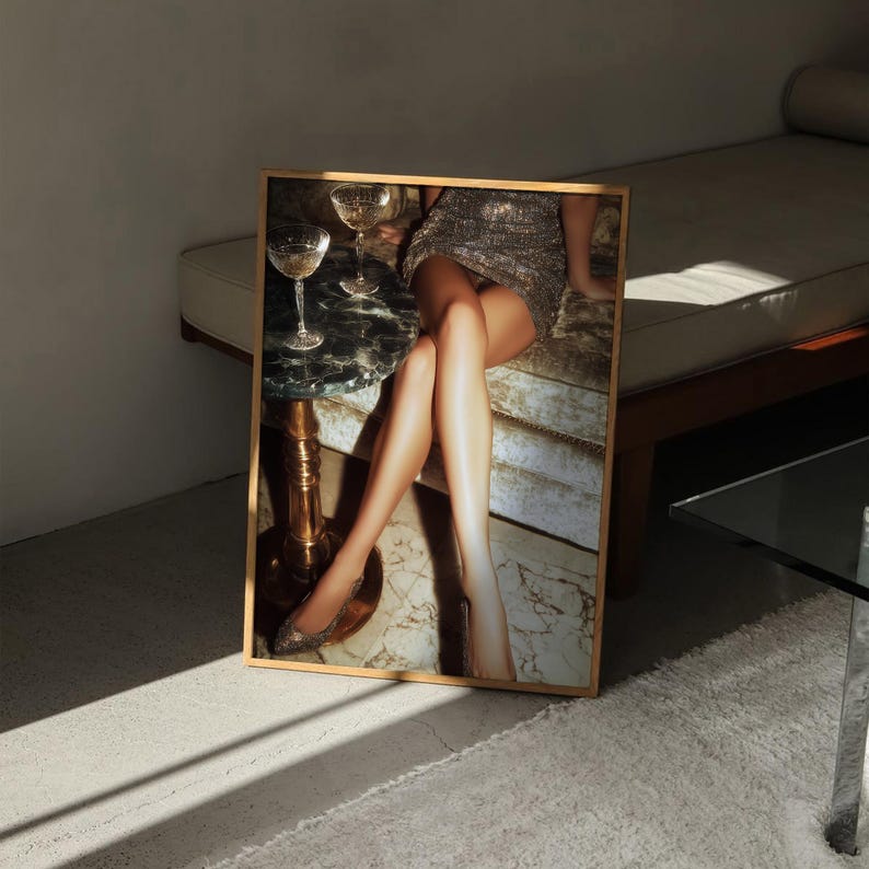 Puede incluir: Impresi&oacute;n art&iacute;stica enmarcada que presenta una escena glamurosa. La imagen muestra a una persona con un vestido plateado brillante, sentada cerca de una peque&ntilde;a mesa con copas de champ&aacute;n. La atenci&oacute;n se centra en las piernas y el ambiente elegante general.