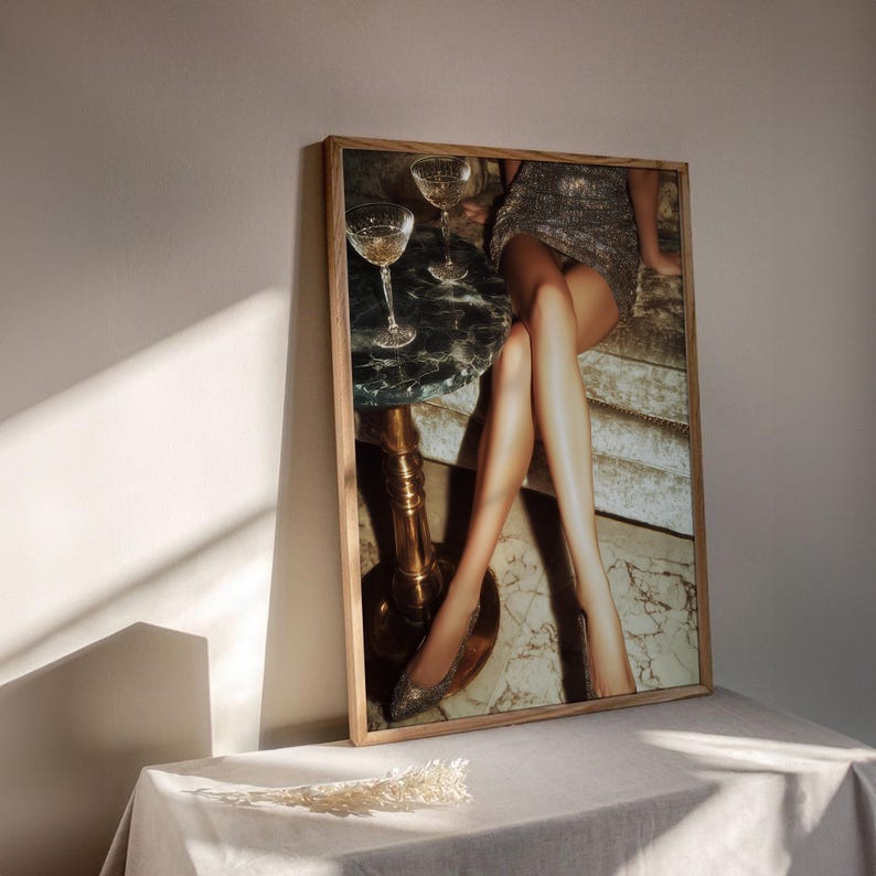 Puede incluir: Una impresi&oacute;n enmarcada que presenta una escena glamurosa. La imagen muestra a una persona con un vestido plateado con lentejuelas, sentada cerca de una peque&ntilde;a mesa de m&aacute;rmol con dos copas de champ&aacute;n. La atenci&oacute;n se centra en las piernas de la persona y el entorno elegante.