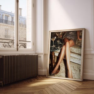 Puede incluir: Una impresi&oacute;n enmarcada se apoya contra una pared, representando una escena glamurosa. La imagen muestra a una persona con un vestido brillante, sentada cerca de una mesa con copas de champ&aacute;n. La atenci&oacute;n se centra en las piernas y la est&eacute;tica lujosa.