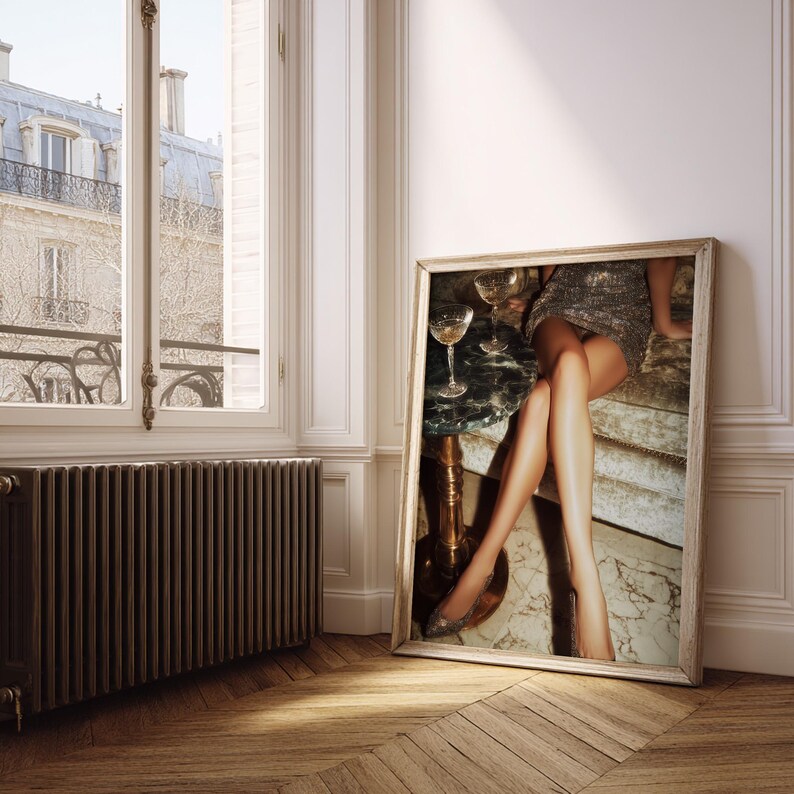 Puede incluir: Una impresi&oacute;n enmarcada se apoya contra una pared, representando una escena glamurosa. La imagen muestra a una persona con un vestido brillante, sentada cerca de una mesa con copas de champ&aacute;n. La atenci&oacute;n se centra en las piernas y la est&eacute;tica lujosa.