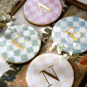 May include: Set of four decorative ceramic coasters with gold trim. Each coaster features a different pastel checkered pattern in pink, green, blue, and beige, with a gold letter in the center. The letters are A, E, E, and N.