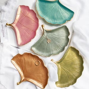 May include: Five decorative ceramic trays shaped like ginkgo leaves. The trays are in various colors, including pink, green, gold, and light green. Each tray has a gold rim and a raised design.