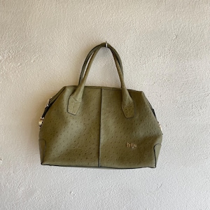 May include: An olive green handbag with a textured finish and two handles. The bag has a structured, rectangular shape and gold-coloured hardware. The brand name "frija" is visible in gold lettering on the front.