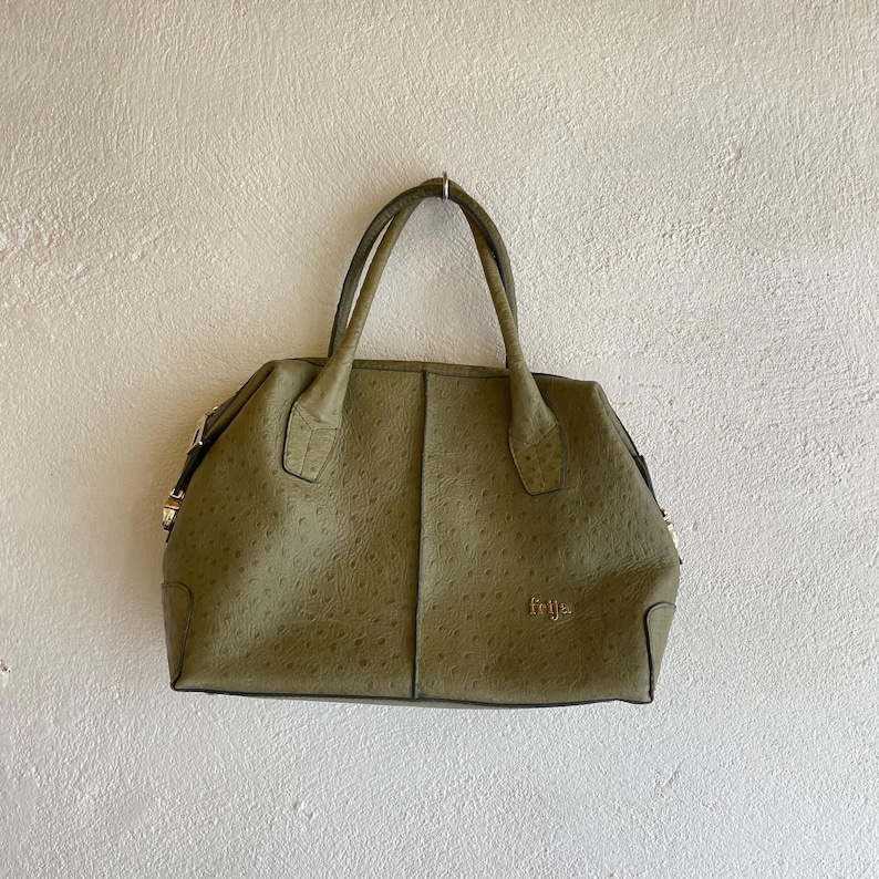 May include: An olive green handbag with a textured finish and two handles. The bag has a structured, rectangular shape and gold-coloured hardware. The brand name "frija" is visible in gold lettering on the front.