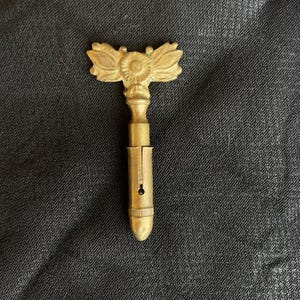 May include: A vintage brass key with a decorative floral design on the handle. The key has a T-shaped handle with a detailed sunburst and leaf motif. The key's shaft is cylindrical with a slot and a small hole.