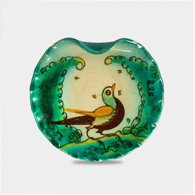 Puede incluir: Un plato redondo de cer&aacute;mica verde con un borde ondulado. El plato est&aacute; decorado con un p&aacute;jaro pintado en tonos de marr&oacute;n, amarillo y verde. El p&aacute;jaro est&aacute; rodeado de espirales y l&iacute;neas verdes.