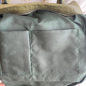 May include: Close-up of the interior of a green leather handbag. The inside features a dark green fabric lining with a pocket. The lining has a repeating pattern. The top of the bag has a zipper closure.