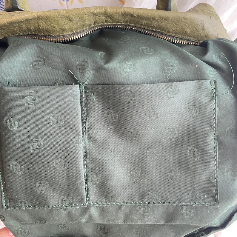 May include: Close-up of the interior of a green leather handbag. The inside features a dark green fabric lining with a pocket. The lining has a repeating pattern. The top of the bag has a zipper closure.