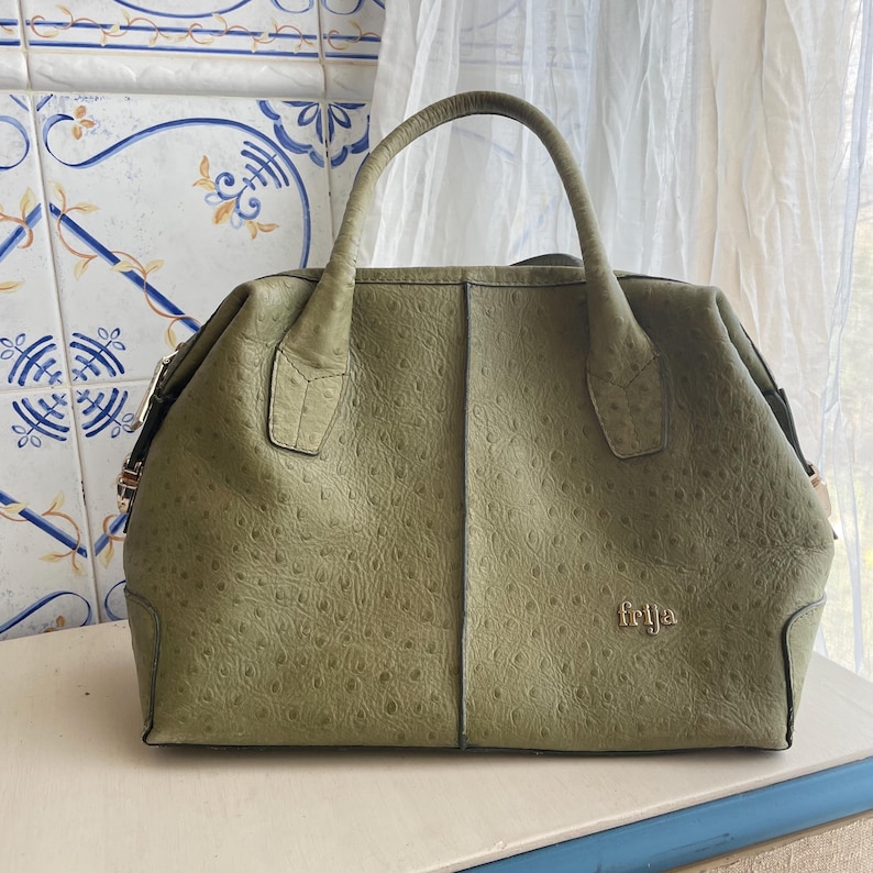 May include: A sage green handbag with a textured, ostrich-like pattern. The bag has a top handle and a gold-coloured clasp. The word "feija" is embossed in gold on the front of the bag.