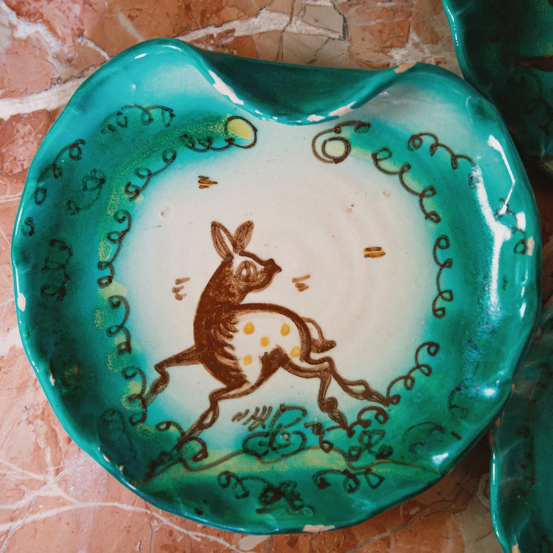 Puede incluir: Un plato de cer&aacute;mica azul turquesa con un centro blanco que presenta un ciervo pintado a mano en marr&oacute;n y blanco con manchas amarillas. El plato tiene un borde ondulado y un borde decorativo de espirales verdes.