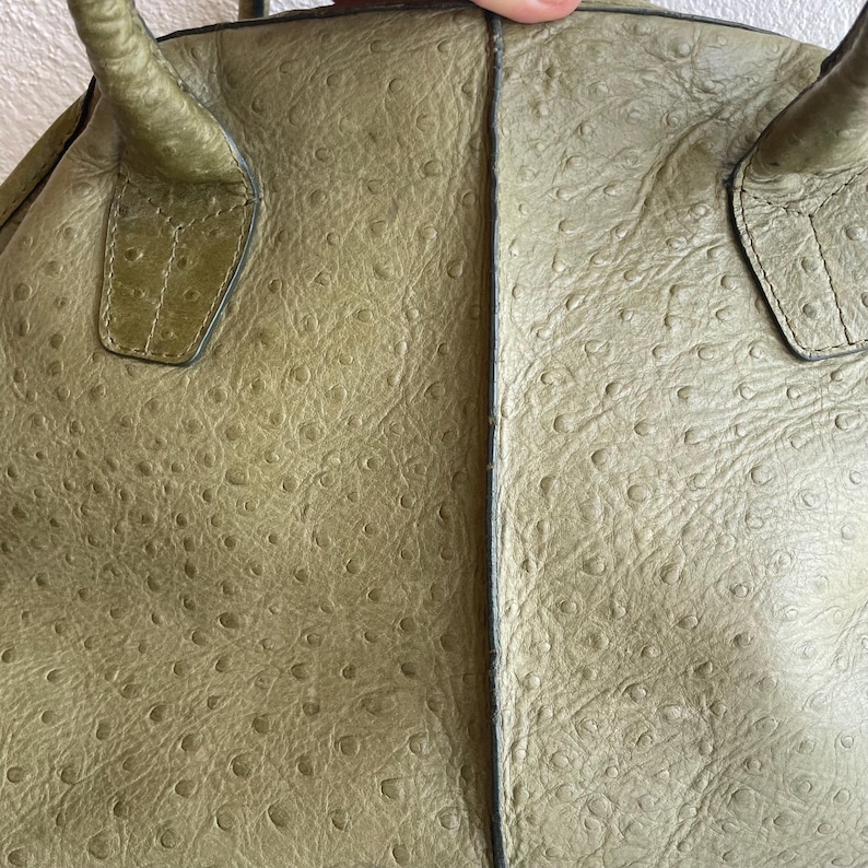 May include: Olive green leather handbag with a textured, dotted pattern. The bag has two handles and a structured design. The leather has a slightly worn appearance, adding to its vintage appeal. The stitching is visible along the seams.