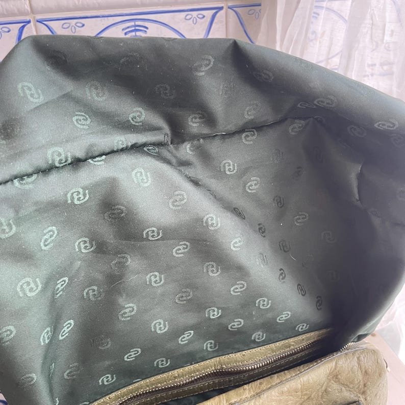 May include: Inside view of a green leather handbag. The lining is a darker green fabric with a repeating pattern of a stylized letter "R". The zip is visible at the bottom of the bag.
