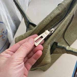 May include: Close-up of a green leather handbag with a textured surface. The bag features a silver zipper and a gold-coloured zipper pull with the brand name "Frija" embossed on it. The bag has two handles.