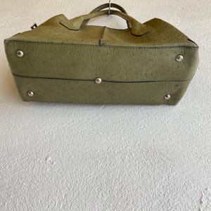 May include: Olive green handbag with a textured surface, silver-tone feet, and a top handle. The bag has a structured shape and appears to be made of leather. The bag is suspended against a white wall.